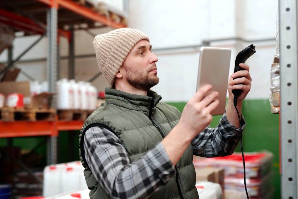 Man in a warehouse managing inventory with a scanner and tablet.