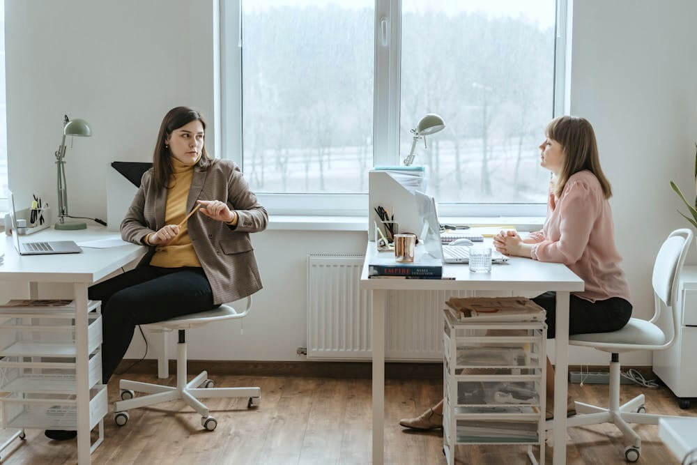 Two women talking to each other behind their desks in office.